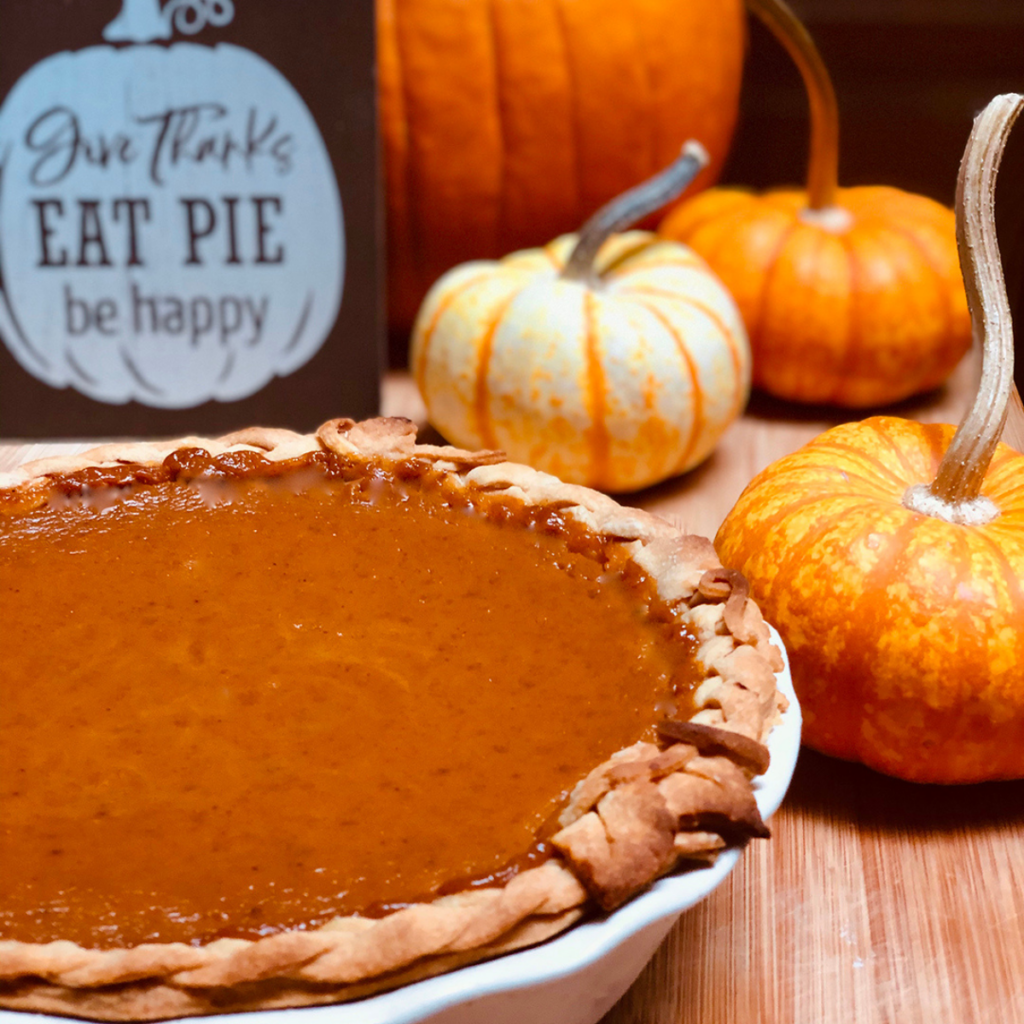A vegan pumpkin pie surrounded ny mini pumpkins and a small wood sign saying "Give thanks, eat pie, be happy.
