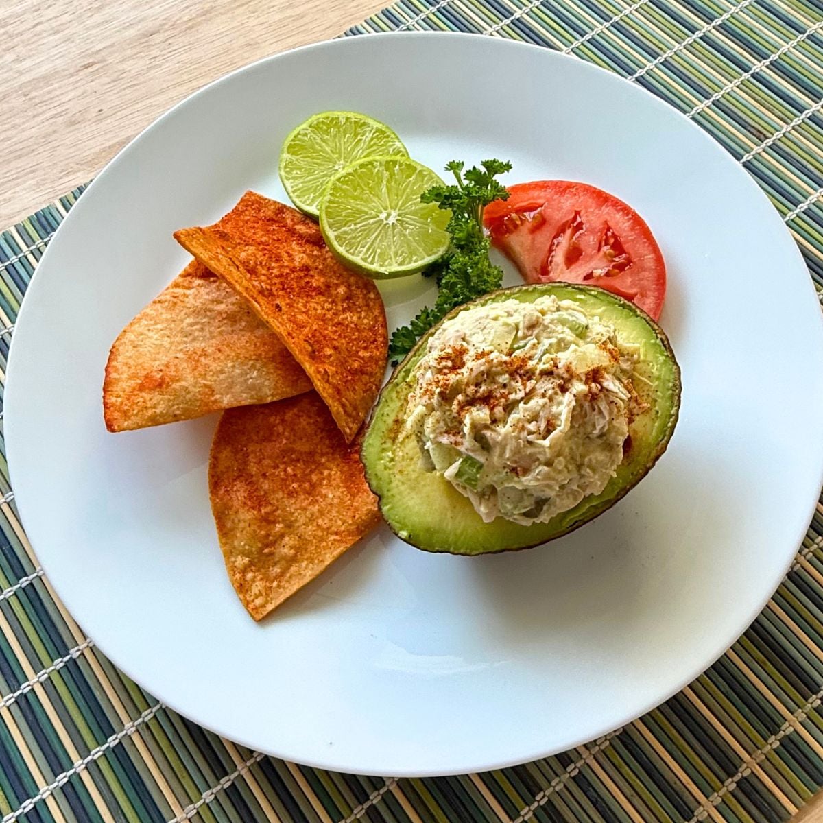 A plate with a half avocado filled with vegan tuna salad surrounded by tortilla chips, lime and tomato slices.