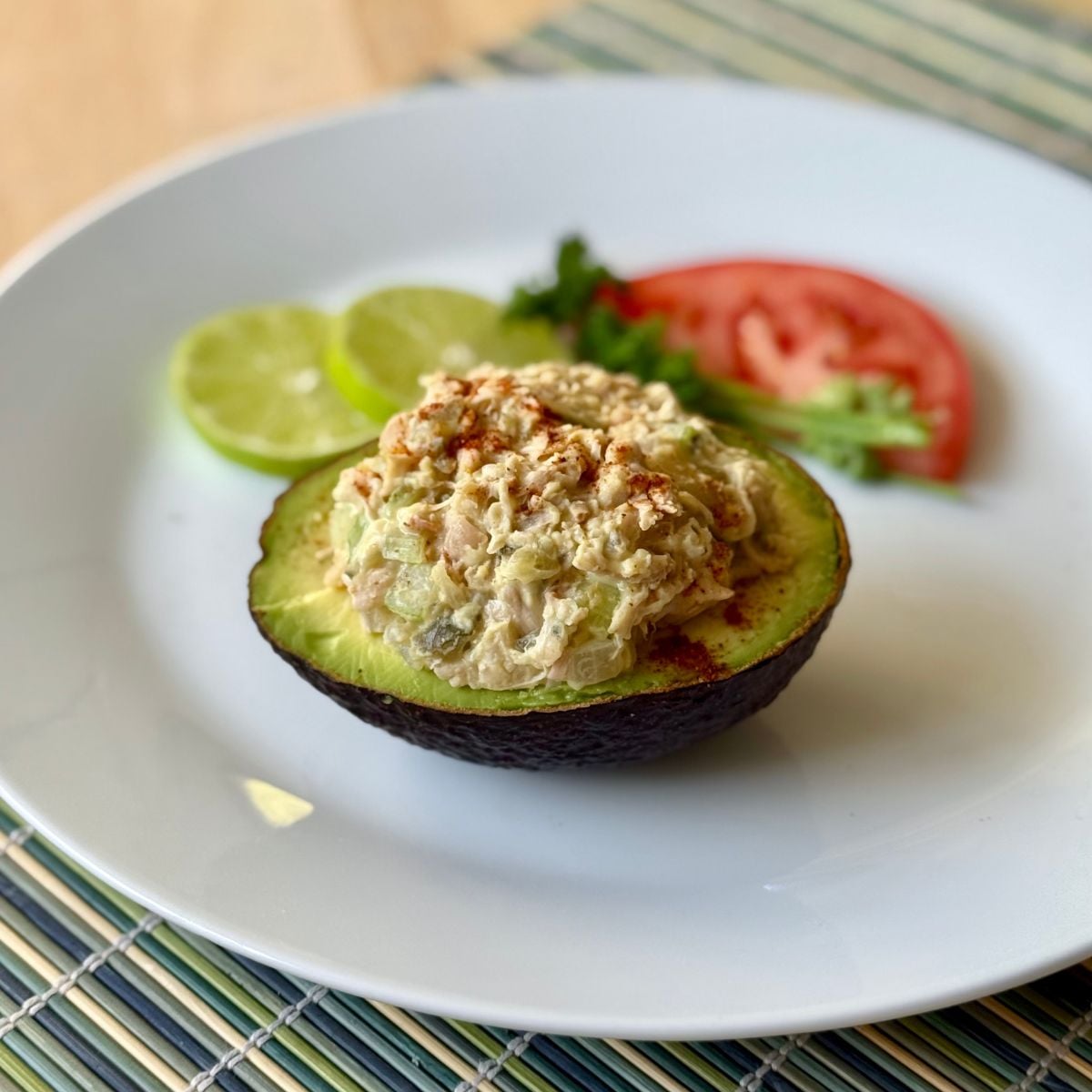 A plate with a half avocado filled with vegan tuna salad surrounded by lime and tomato slices.