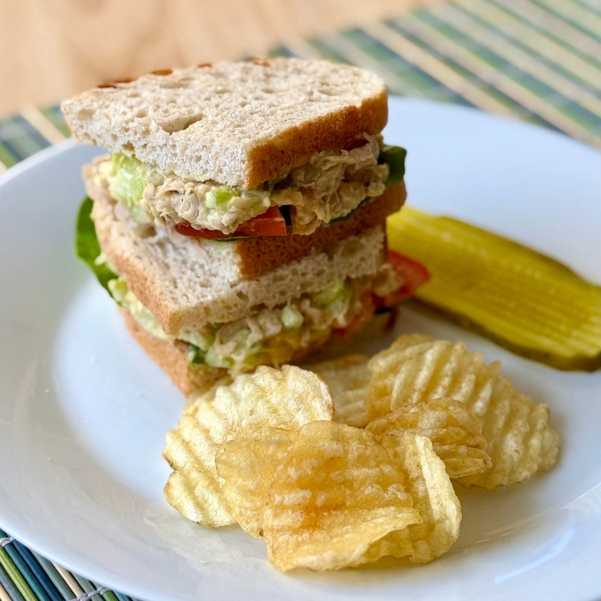 A plate with a stacked vegan tuna salad sandwich, pickle slices and potato chips.