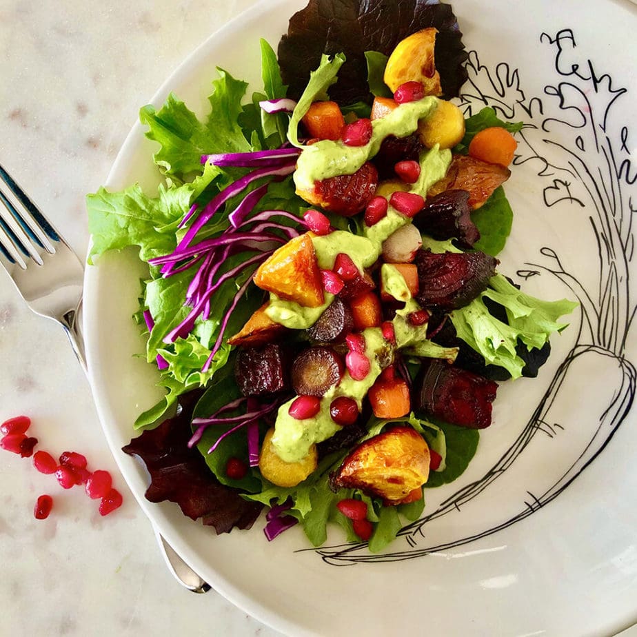 Roasted carrots and beets served on a plate with romaine, red cabbage, vegan avocado basil dressing and pomegranate seeds.