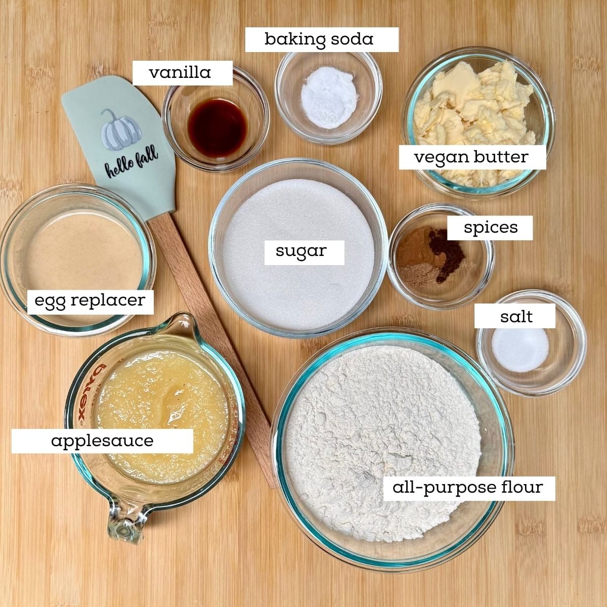 Bowls filled with ingredients for preparing a vegan old-fashioned applesauce cake including: applesauce, flour, sugar, butter, egg-replacer, spices, vanilla, baking soda, and salt.
