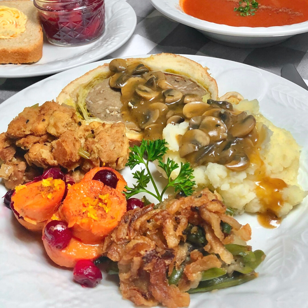 A white dinner plate with a vegan Thanksgiving meal of mushroom seitan Wellington, mashed potatoes and mushroom gravy, bread stuffing, maple cranberry yams and green bean casserole.