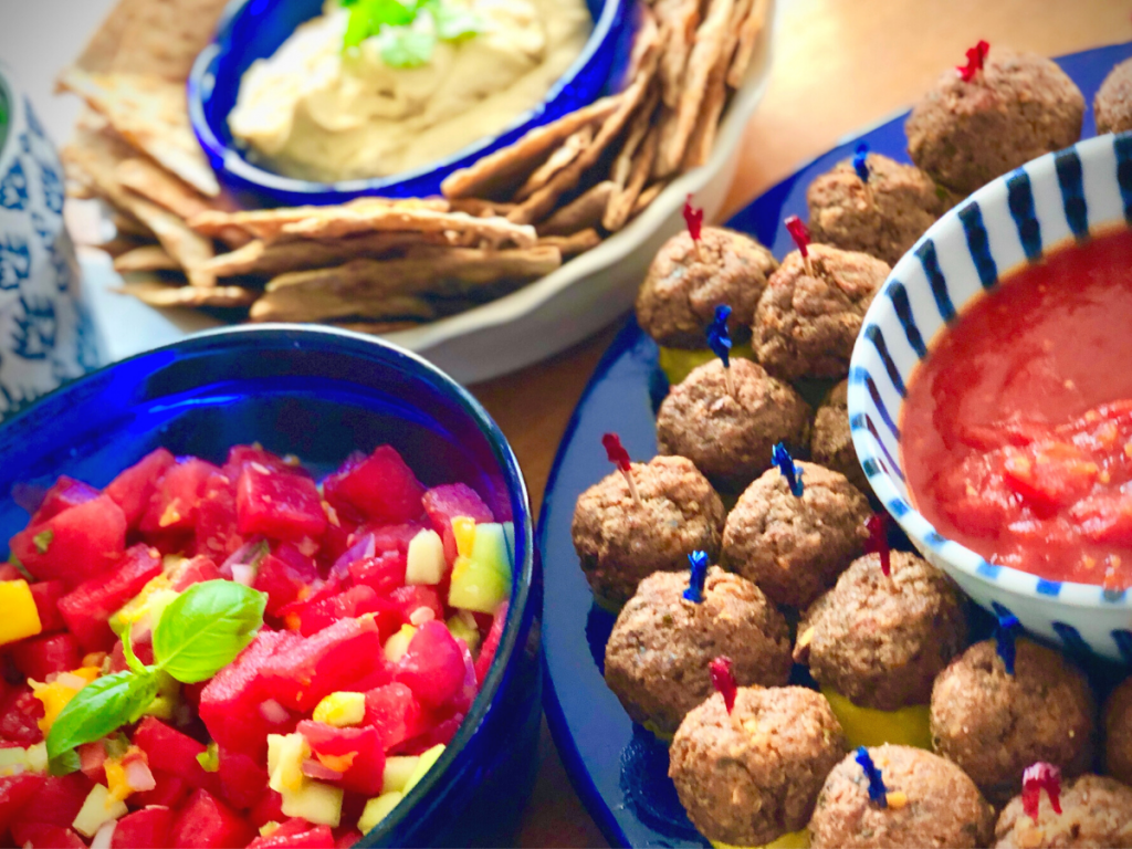 An image of blue bowls with vegan 4th of July foods including vegan meatballs, watermelon salad and hummus with chips.