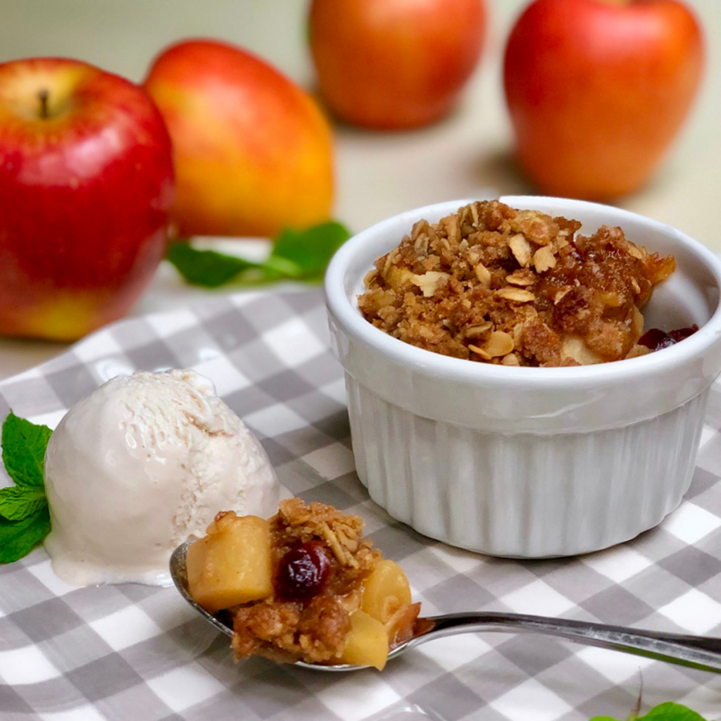 A white ramekin with vegan apple crisp set atop a grey and white checkered plate with a scoop of vegan ice cream. There are red apples in the background.