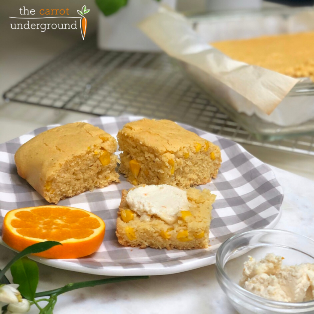 A grey and white gingham ceramic plate with three slices of vegan cornbread and butter and an orange slice.