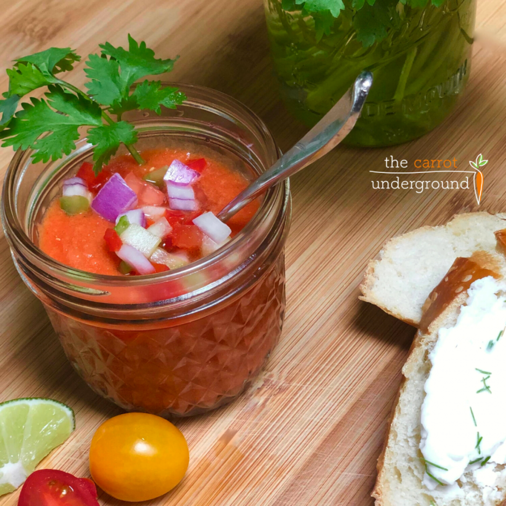 A jam jar filled with chilled gazpacho soup sprinkled with diced cucumber, red onion and bell peppers and a spring of cilantro.