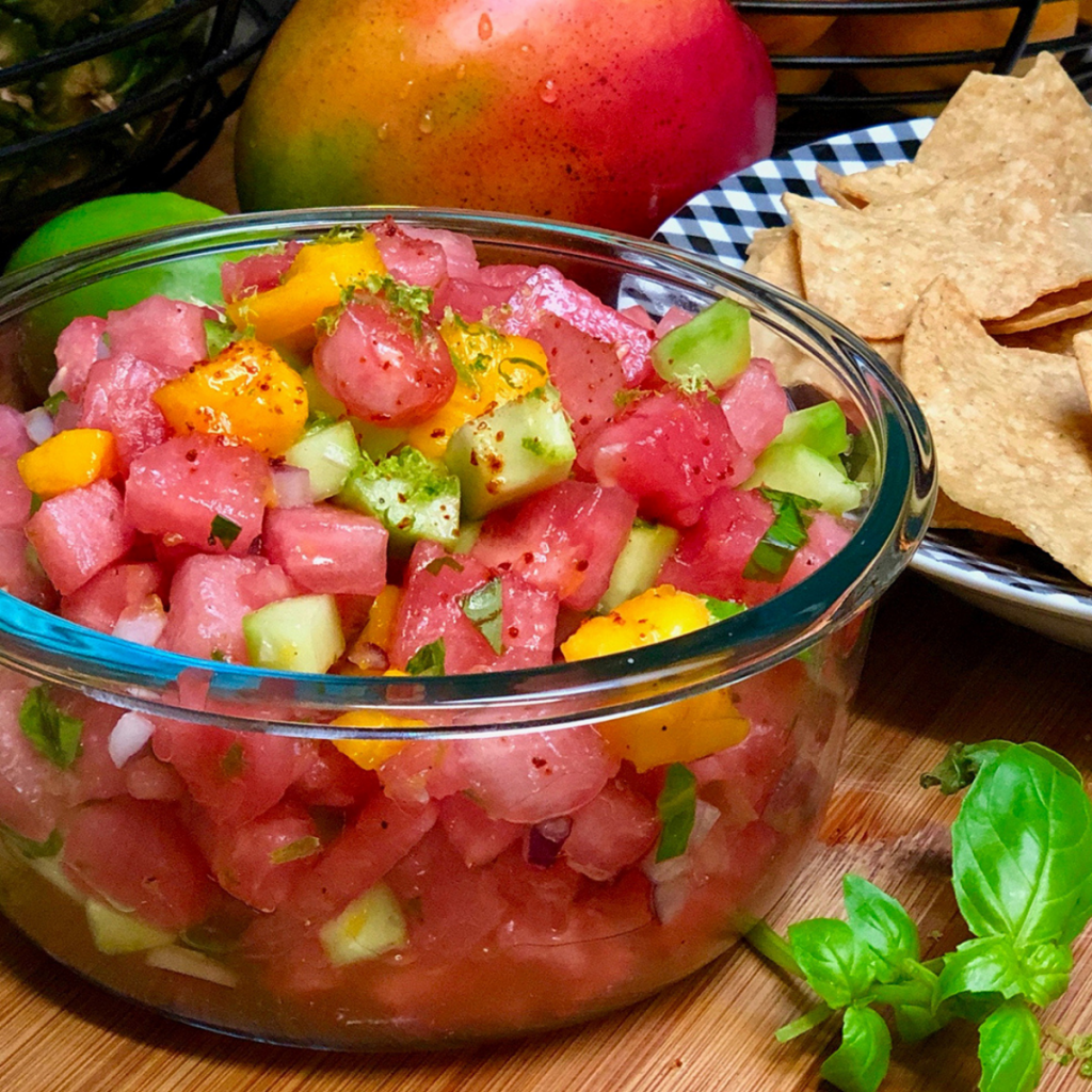 A glass bowl filled with watermelon, mango and cucumber pico de gallo arranged next to a bowl of chips and fresh fruit.