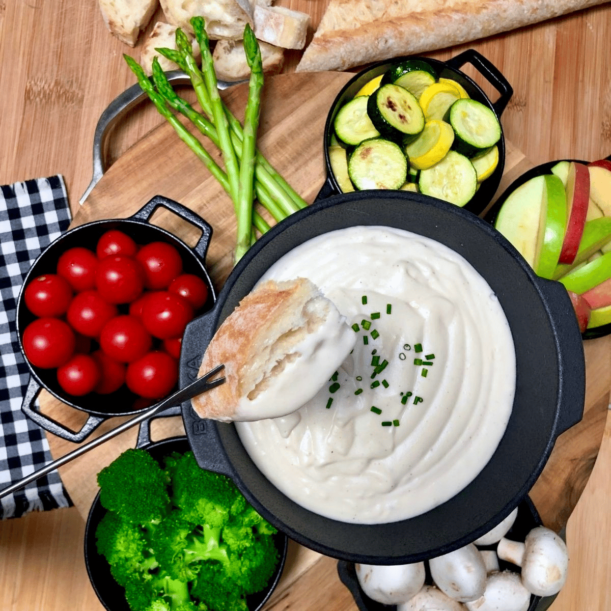 A cast iron fondue pot filled with vegan cheese fondue, surrounded by bowls of sliced apples, broccoli florets, cherry tomatoes, grilled squash, asparagus, and a sliced baguette.