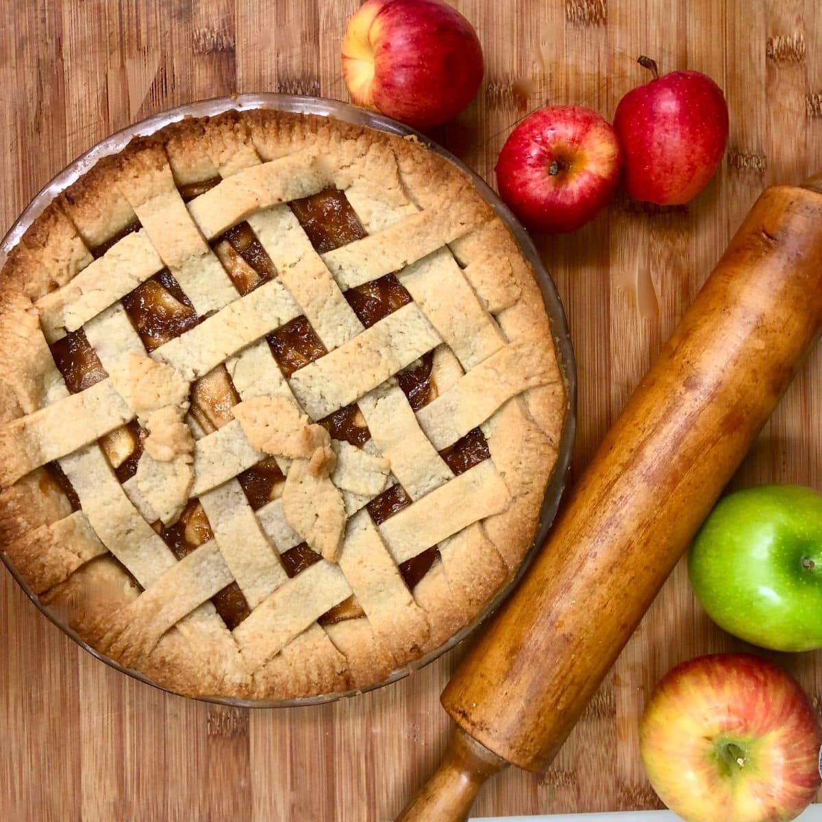 A vegan apple pie topped with a lattice crust, on a cutting board with red and green apples and a rolling pin.