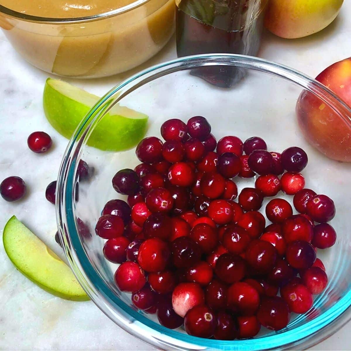 A bowl filled with fresh cranberries, next to apple slices and a dish with applesauce.