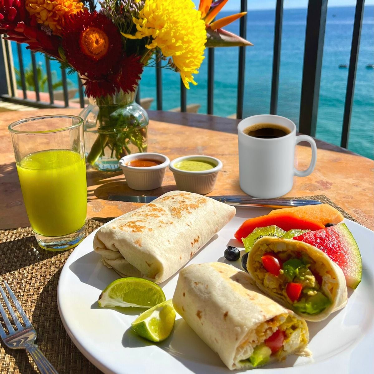 A beachside breakfast table set with a plate of vegan California breakfast burritos, and fruit, along with a mud of coffee, a glass of fresh green juice, and a vase filled with with flowers.