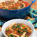 A bowl filled with vegan minestrone soup placed next to an enameled cast iron Dutch oven filled with soup.