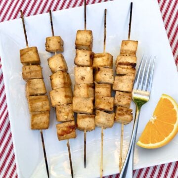 Five bamboo skewers holding grilled tofu with citrus marinade and an orange slice on a plate with a fork.