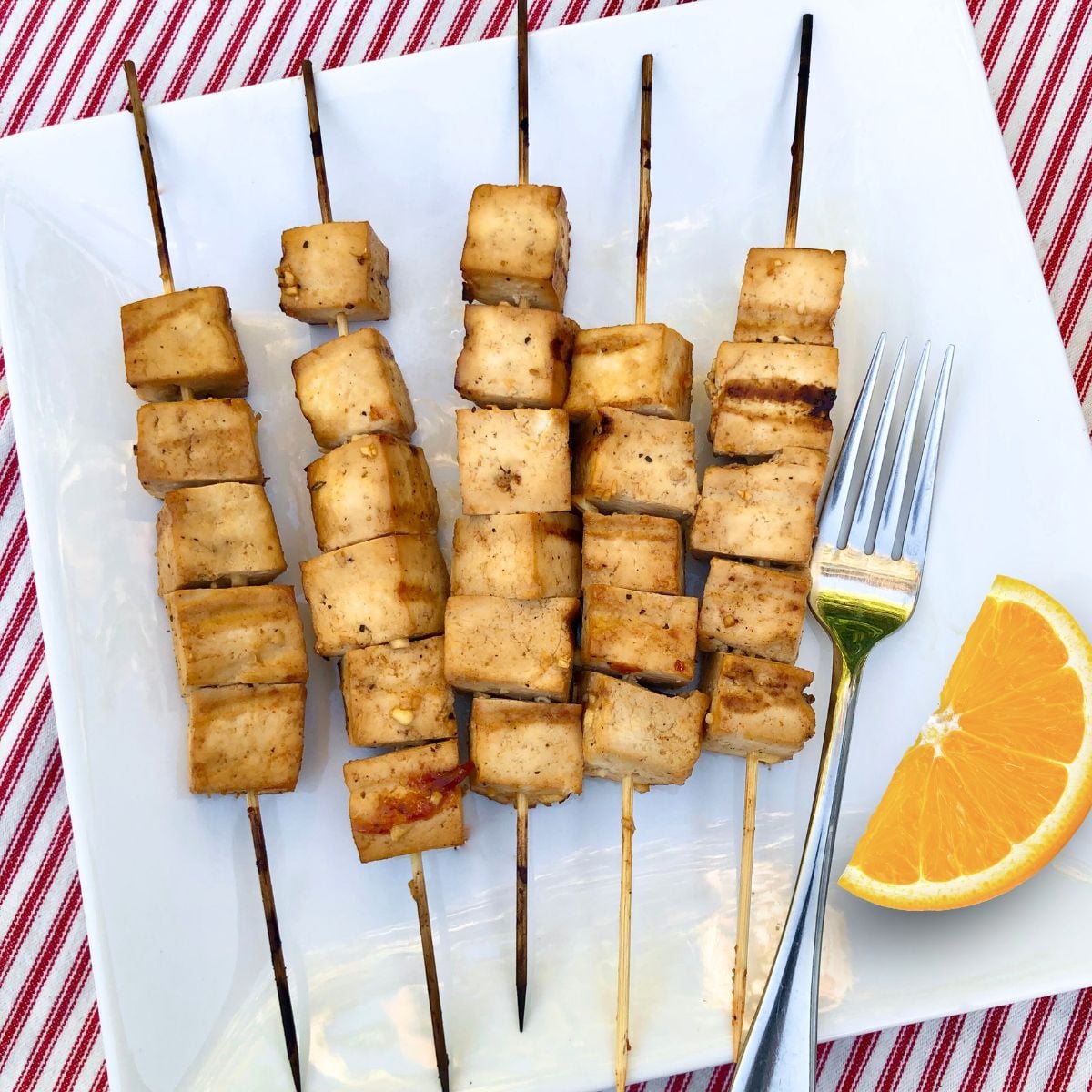Five bamboo skewers holding grilled tofu with citrus marinade and an orange slice on a plate with a fork.
