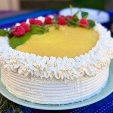 A vegan lemon cake frosted with lemon cream cheese frosting and topped with lemon curd and decorated with fresh raspberries.