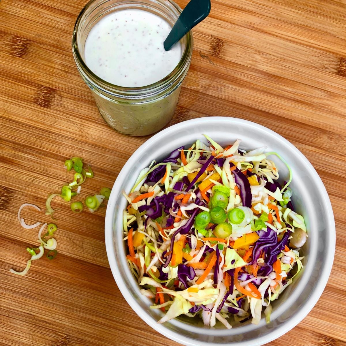 A bowl of shredded cabbage, carrots, and sliced green onions next to a jar filled with classic vegan coleslaw dressing.