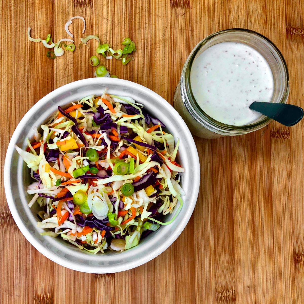 A bowl of shredded cabbage, carrots, and sliced green onions next to a jar filled with classic vegan coleslaw dressing.