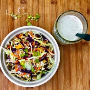 A bowl of shredded cabbage, carrots, and sliced green onions next to a jar filled with classic vegan coleslaw dressing.