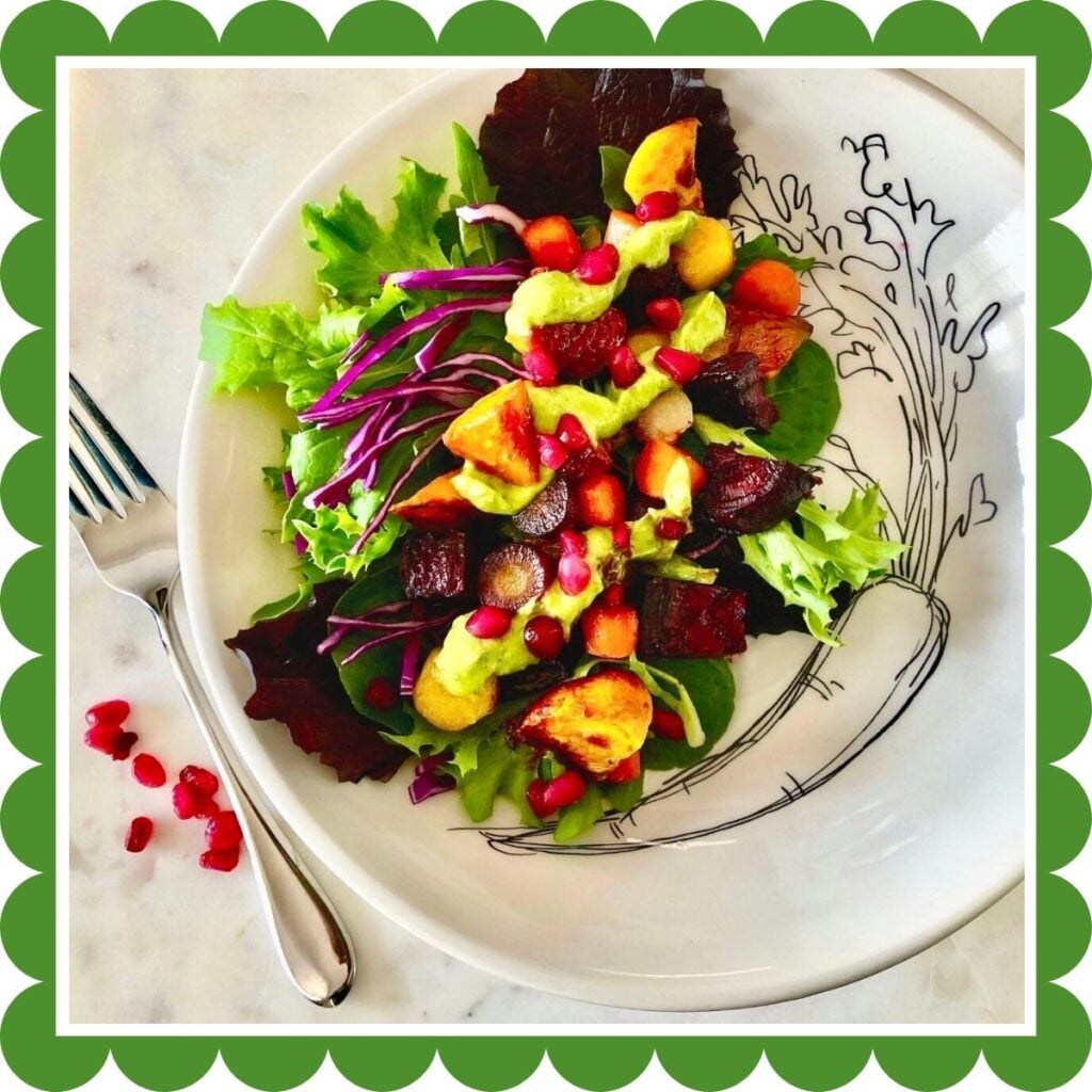 A bowl with roasted root vegetables on a bed of salad greens, topped with avocado basil dressing and pomegranate seeds.