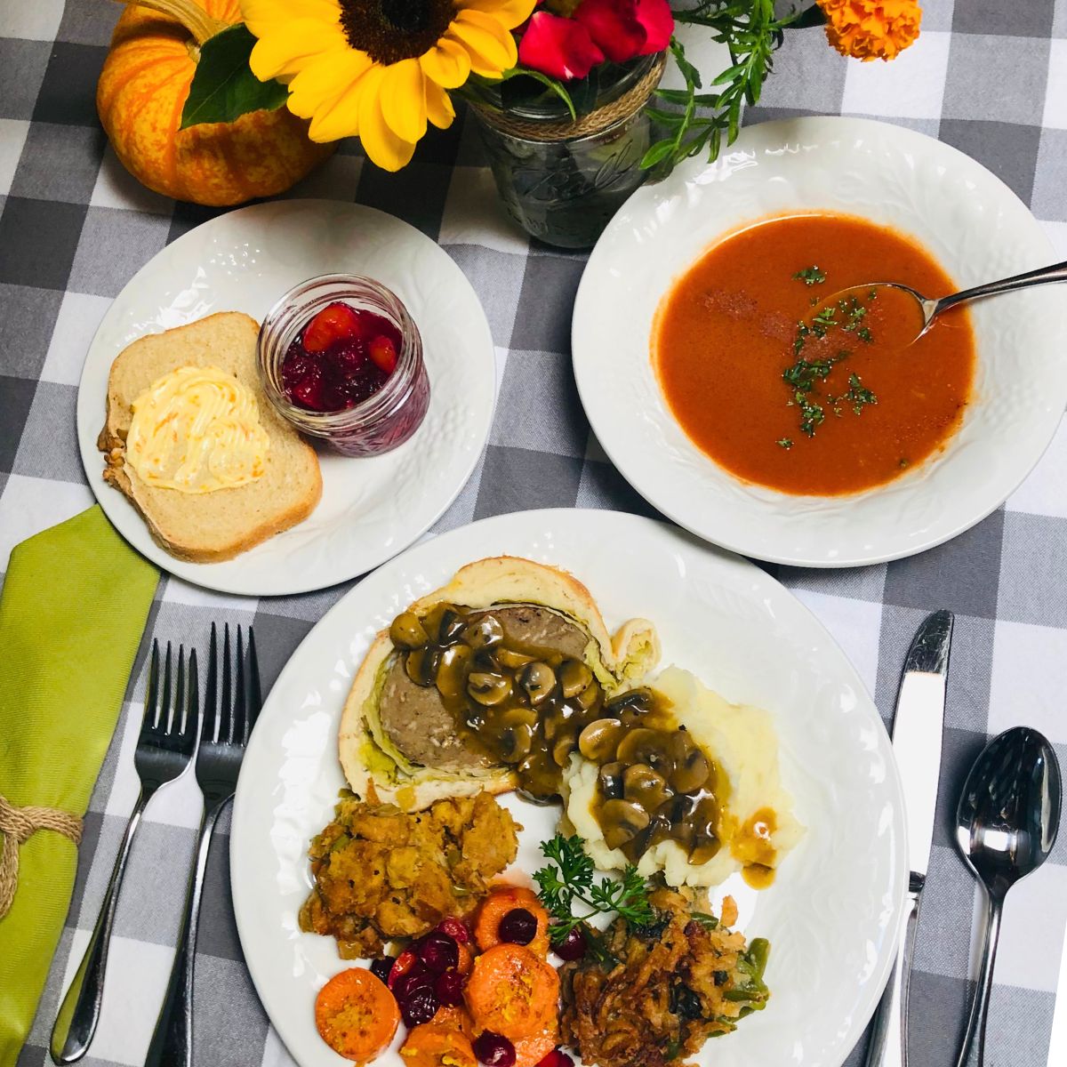 A vegan Thanksgiving dinner plate with a slice of mushroom seitan Wellington, mashed potatoes with mushroom gravy, dressing, classic green bean casserole, cranberry yams, and a bowl of homemade tomato soup, fresh baked bread and cranberry sauce.