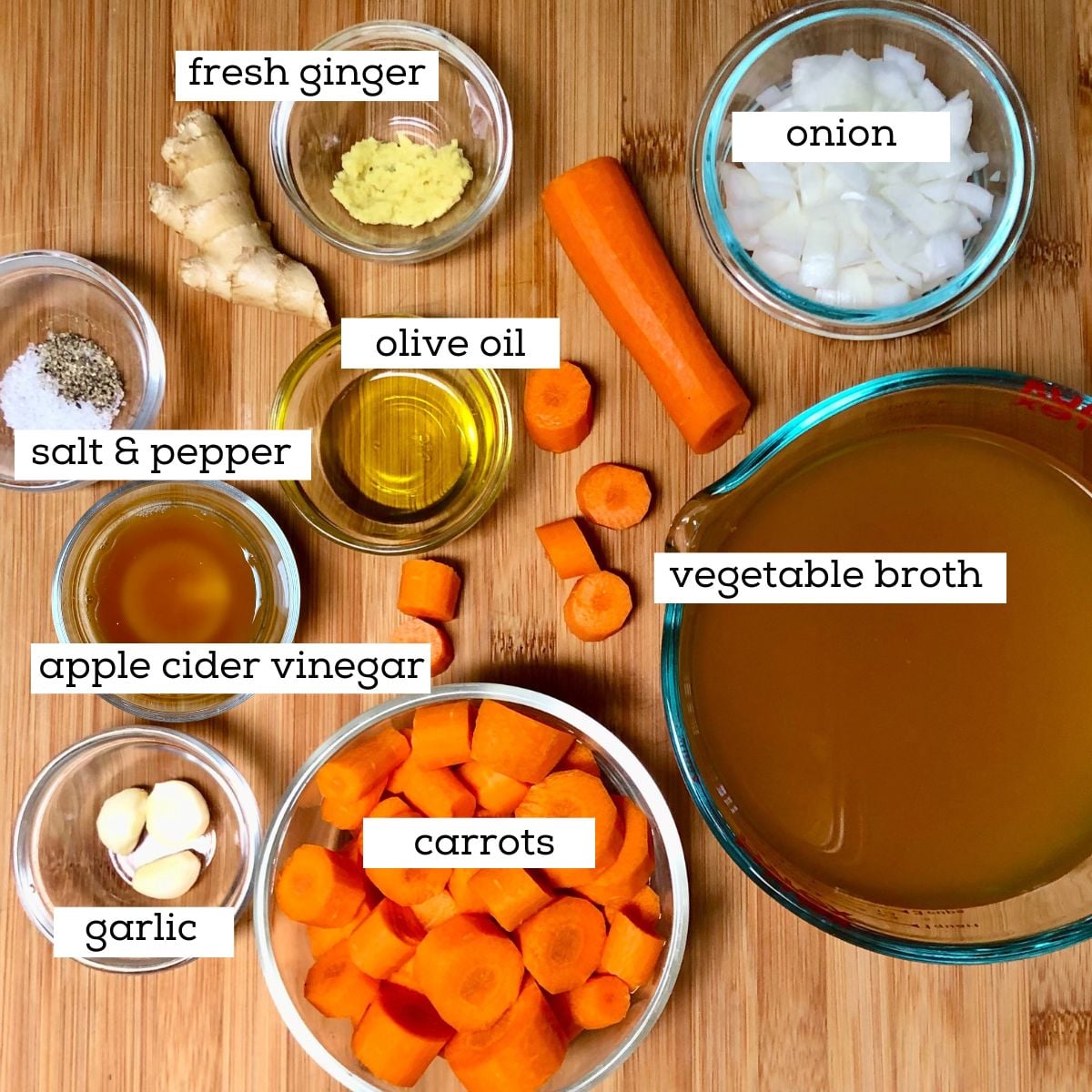 Ingredients for vegan carrot ginger soup in glass bowls, including carrots, ginger, onion, broth, olive oil, garlic, fresh lemon, salt & pepper.