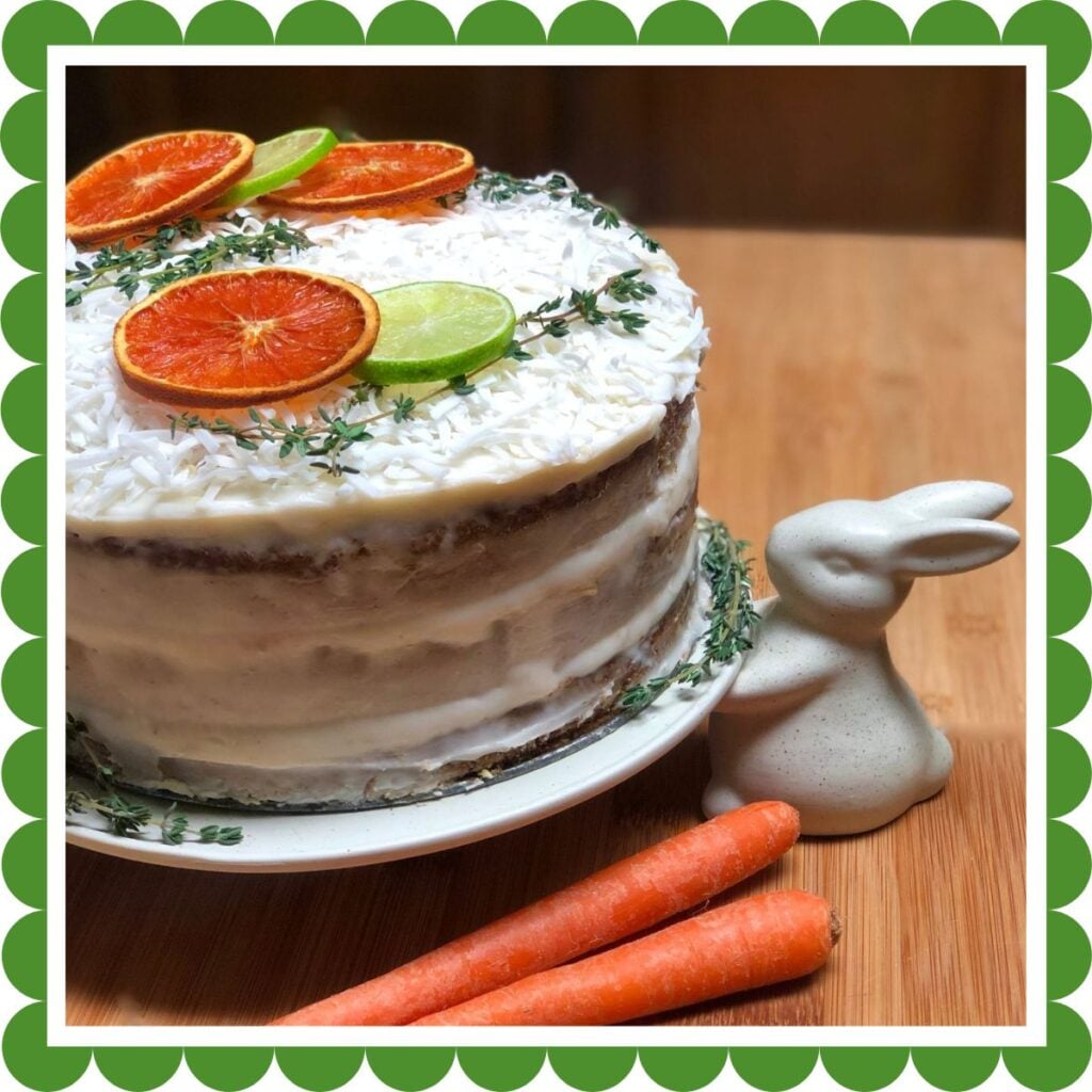 A vegan pineapple carrot cake decorated with cream cheese frosting topped with a sprinkle of flaked coconut and slices of dried orange and lime on a cake stand with a ceramic rabbit holding the plate.