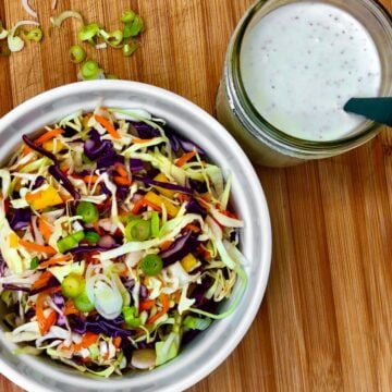 A bowl of shredded cabbage, carrots, and sliced green onions next to a jar filled with classic vegan coleslaw dressing.