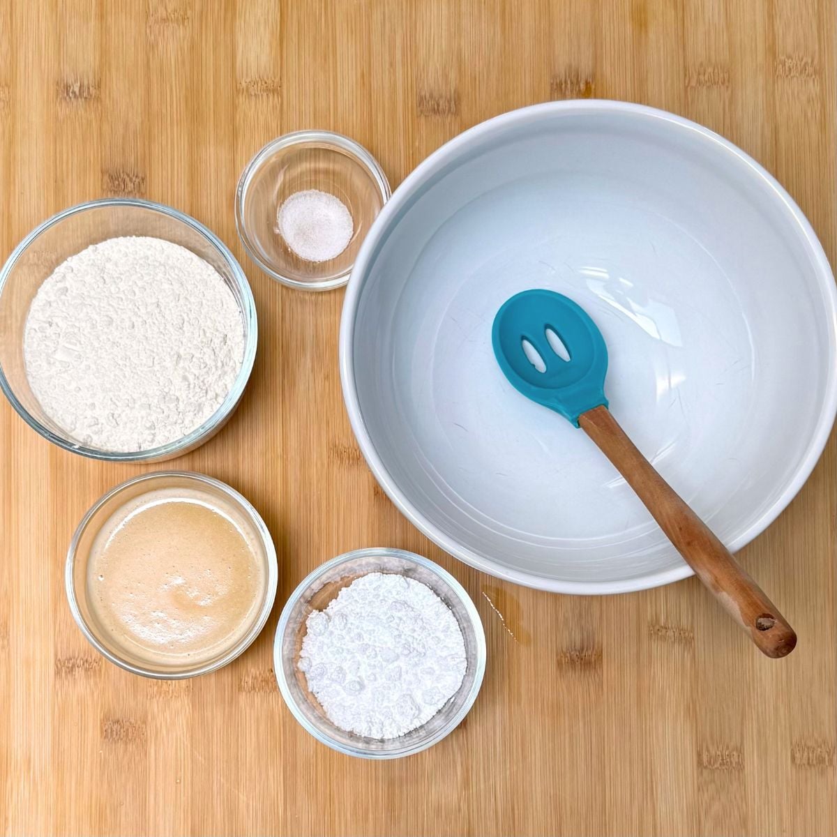 A mixing bowl with spatula next to small bowls with flour, salt, powdered sugar, and melted vegan butter.