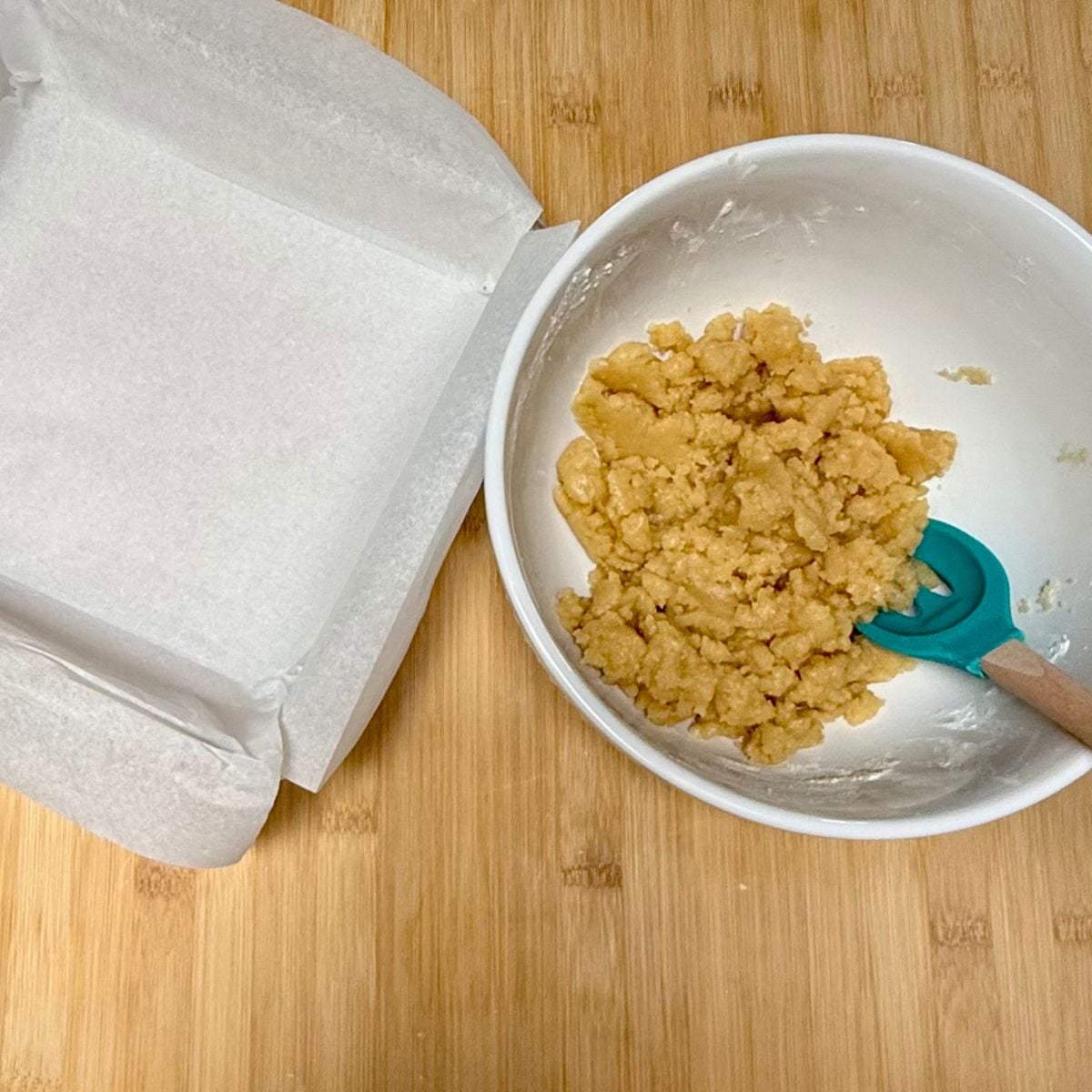 A parchment lined square baking dish next to a bowl with shortbread dough.