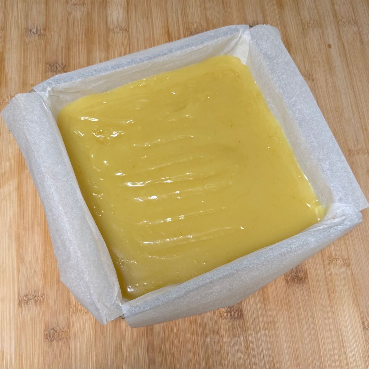 A square parchment lined baking dish with a baked shortbread crust and lemon filling on top.