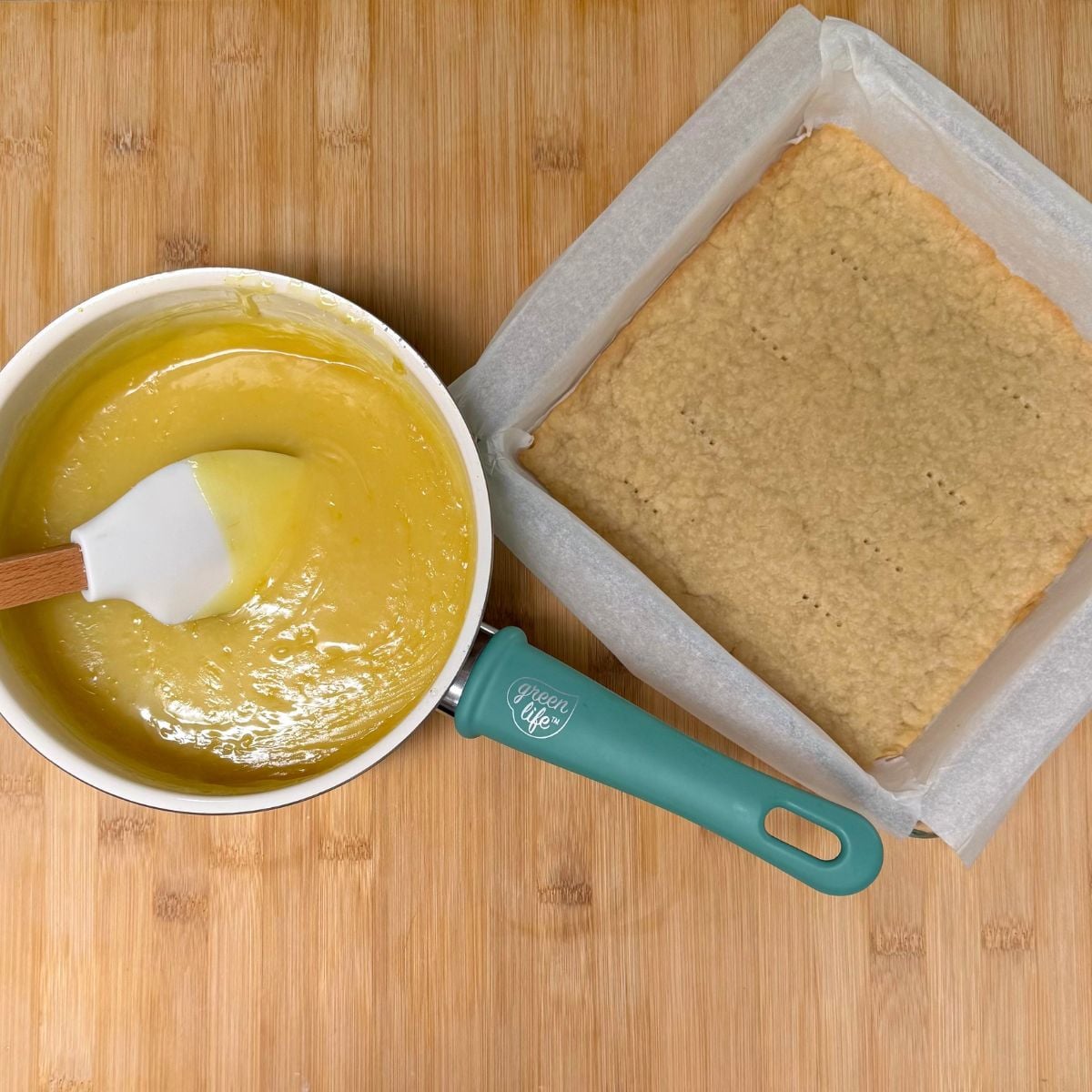 A pre-baked shortbread crust in a square parchment lined baking dish next to a saucepan filled with vegan lemon bar filling.