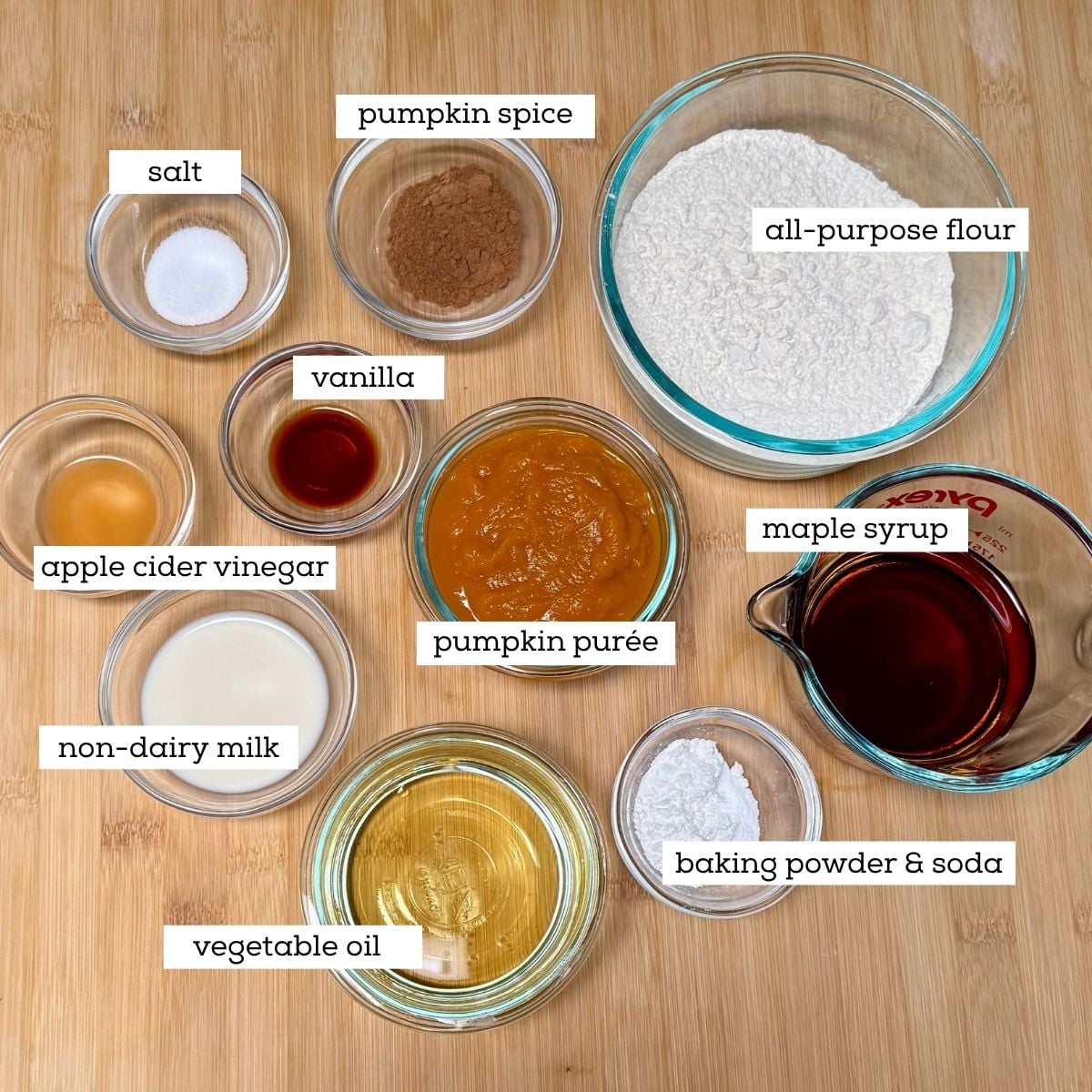 Bowls filled with ingredients for making vegan maple pumpkin muffins including: pumpkin purée, maple syrup, flour, baking powder and soda, pumpkin spice, salt, vanilla, non-dairy milk, apple cider vinegar, and vegetable oil.