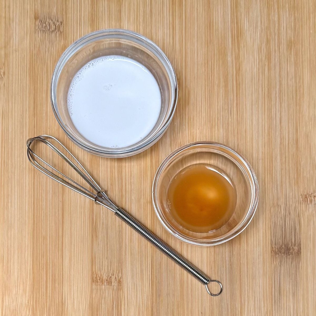 A small bowl with soy milk and another with apple cider vinegar next to a wire whisk.