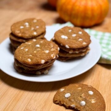 Three stacks of vegan pumpkin white chocolate chip cookies on a plate, and a half eaten cookie, napkin, and mini pumpkin on a table top.