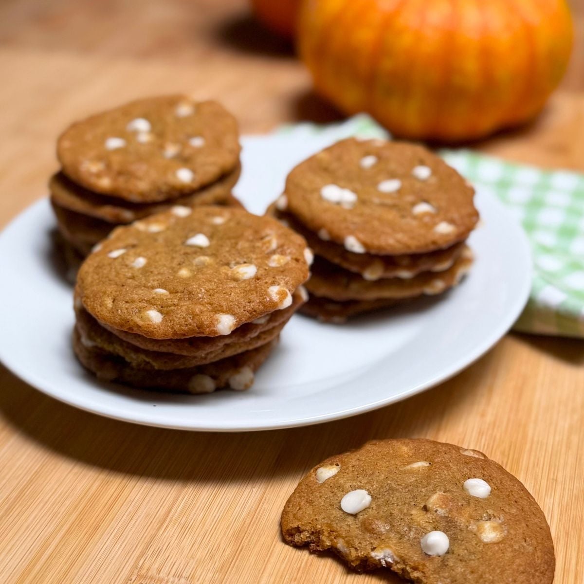 Three stacks of vegan pumpkin white chocolate chip cookies on a plate, and a half eaten cookie, napkin, and mini pumpkin on a table top.