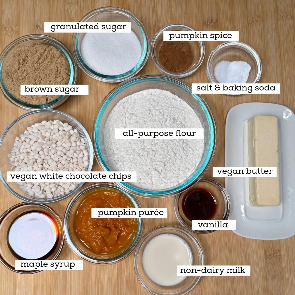 Bowls filled with ingredients for making Vegan Pumpkin Cookies with White Chocolate Chips including, pumpkin purée, white chocolate chips, all-purpose flour, white and brown sugar, vegan butter, maple syrup, vanilla extract, non-dairy milk, baking soda and salt, and pumpkin spice.