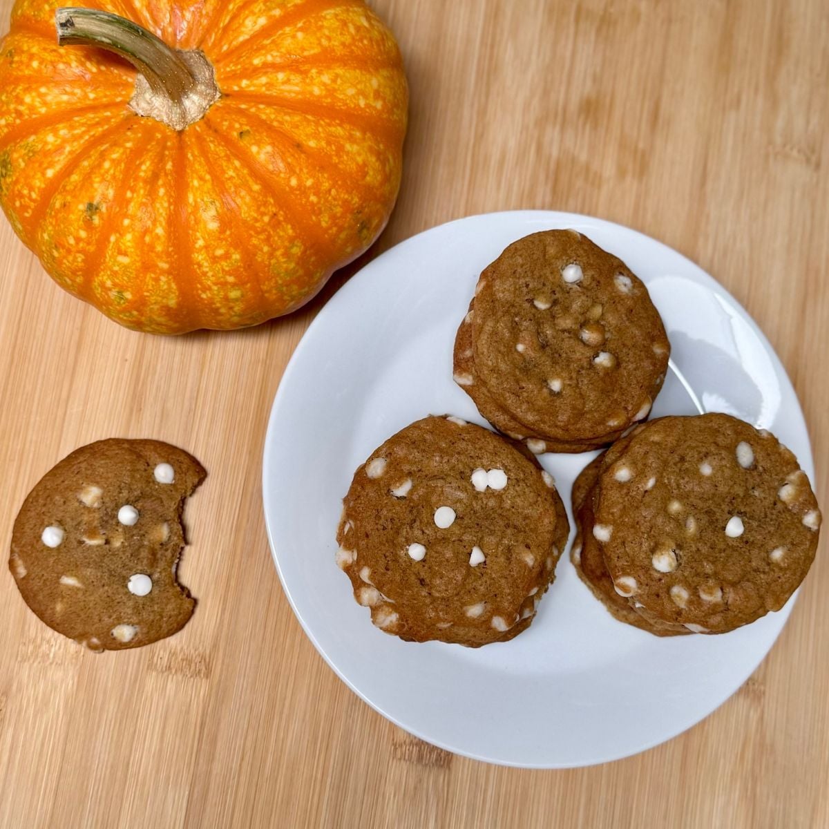 Three stacks of vegan pumpkin white chocolate chip cookies on a plate, and a half eaten cookie, and mini pumpkin on a table top.