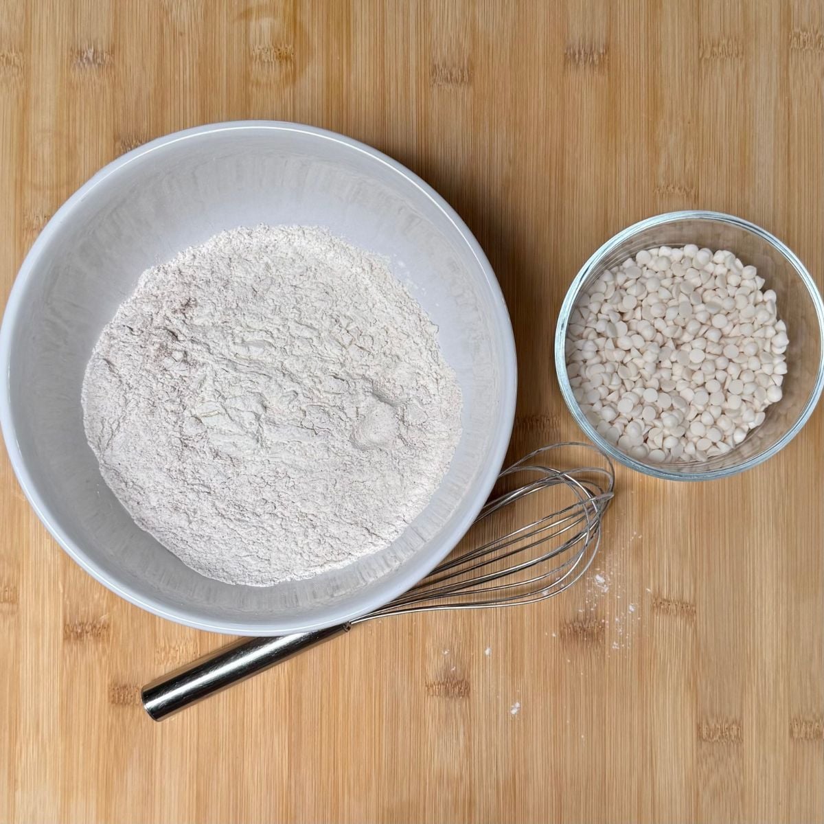 Two bowls on a bamboo cutting board. One filled with dry ingredients for vegan pumpkin cookies with white chocolate chips, the other filled with white chocolate chips.